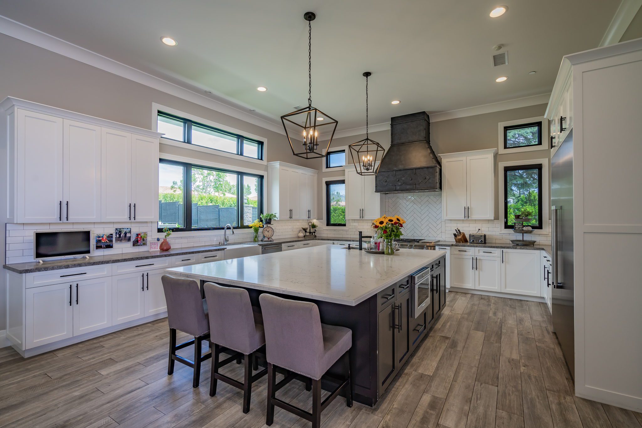 A modern kitchen with white cabinets, a large island with gray chairs, stainless steel appliances, and pendant lighting. Bright windows illuminate the space.